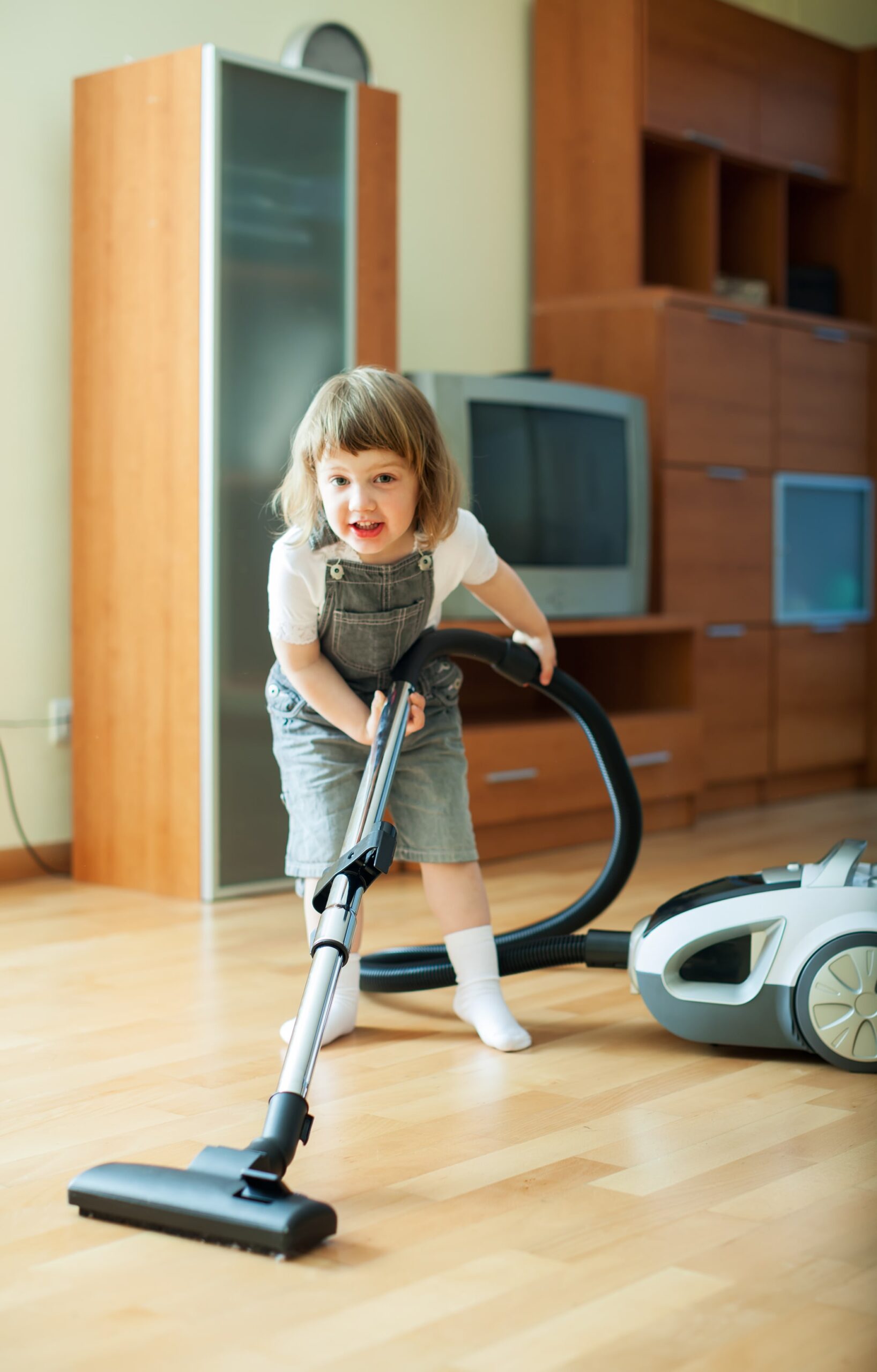 Child in overalls struggling to use a vacuum cleaner, highlighting playful engagement with household chores in a sunny living room.
