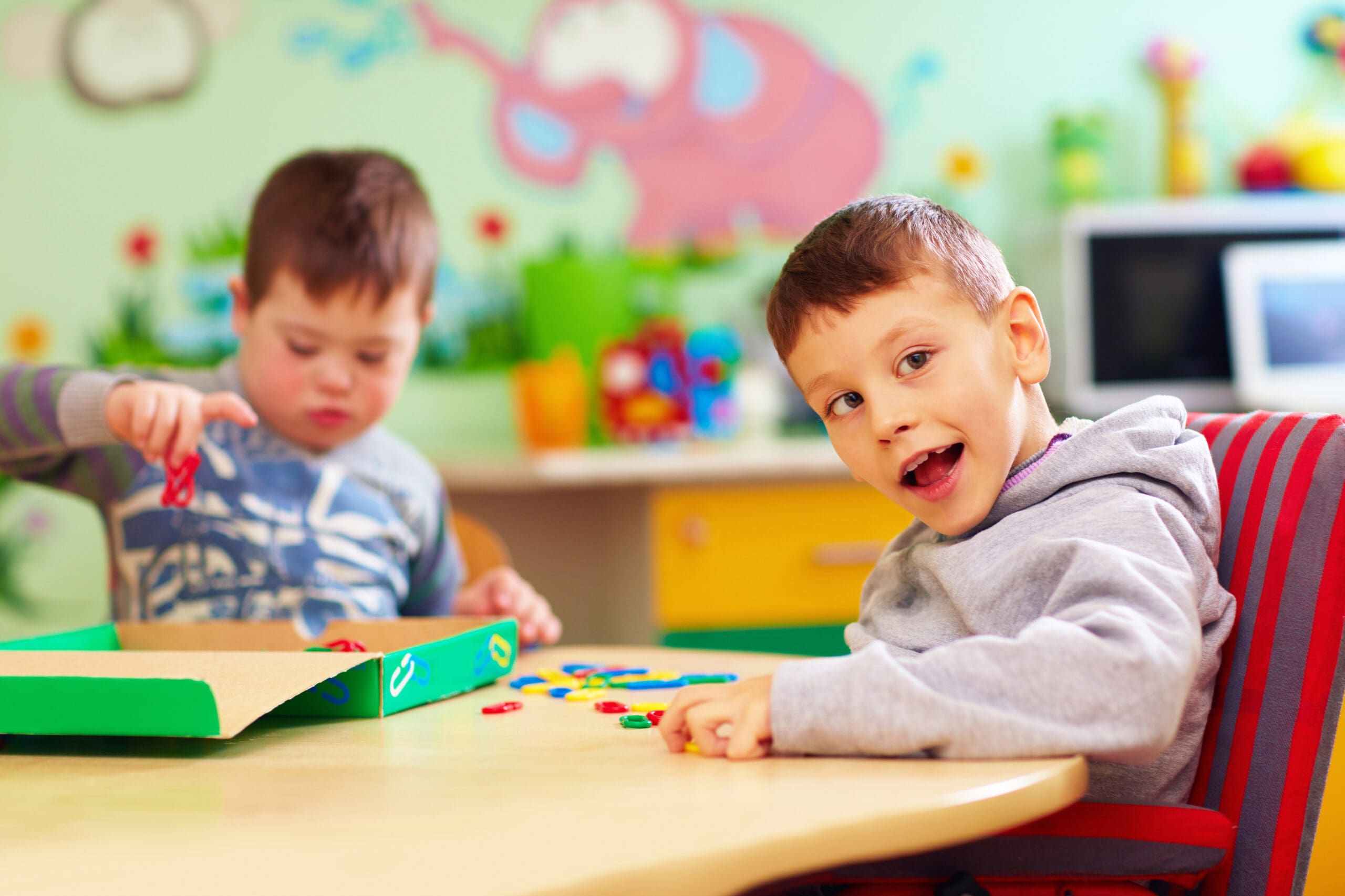 Two children are engaged in play at a colorful table. One child looks happily at the camera while the other focuses on selecting colorful pieces from a box.